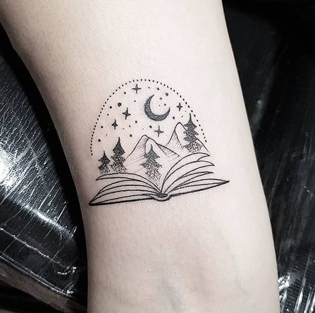 Livre ouvert avec tatouage croissant de lune, étoiles, arbres et montagnes.